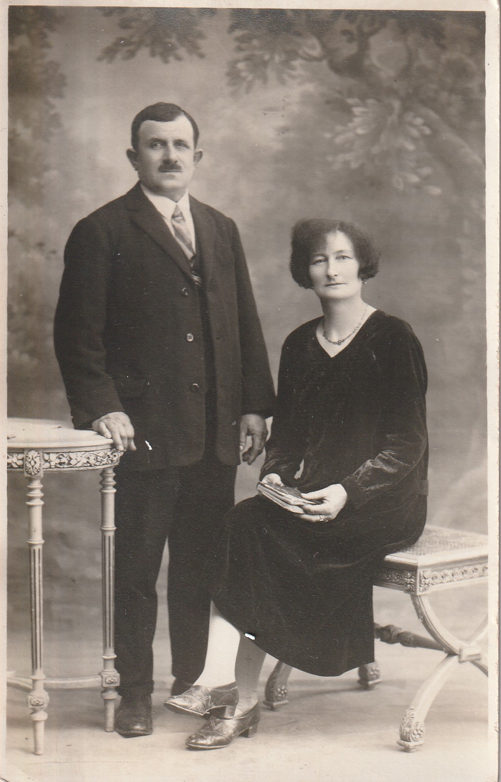 Une carte postale de 1900 qui montre un couple qui pose dans un studio photo
       devant un décor artificiel; une table et une chaise Louis XVI; la femme est assise,
      elle porte une robe sobre et tient un livre dans ses mains; l'homme est
      debout en costume, il a une attitude dominatrice de vieux mâle.