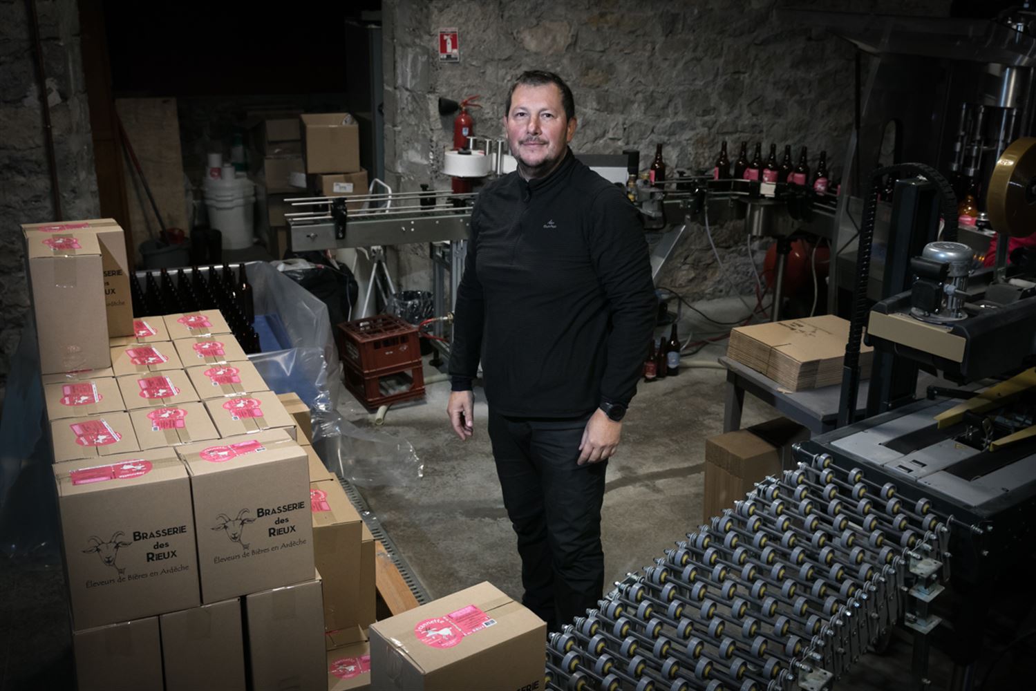 Dans sa brasserie, Florent pose fièrement au milieu de sa chaîne d'embouteillage, parmi cartons emballés et bouteilles encore vides.
