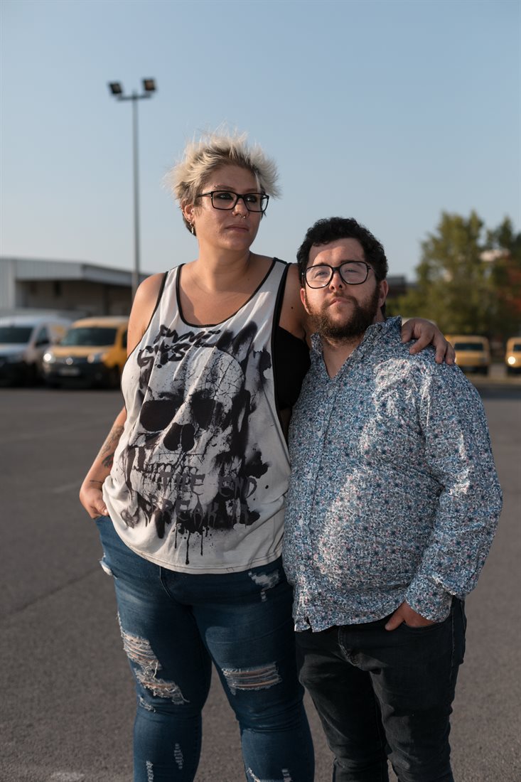 Raphaël et Audrey se tiennent par la taille, sur un parking presque vide, leur visage baigné par la lumière dorée du soleil couchant. 