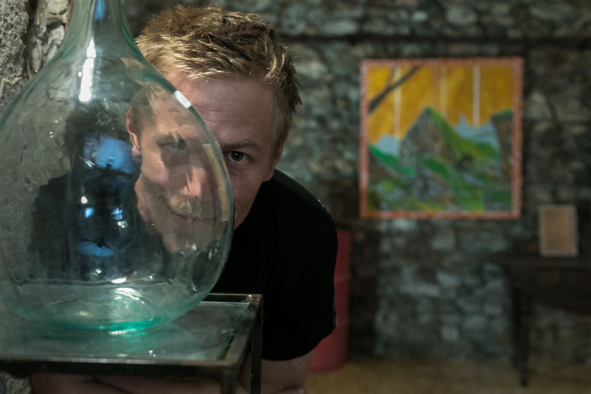 Jérémy, Hilarone, dessinateur, visage demi-caché derrière grosse dame-jeanne vide et transparente, salle d'exposition, en fond une de ses oeuvres orange et verte