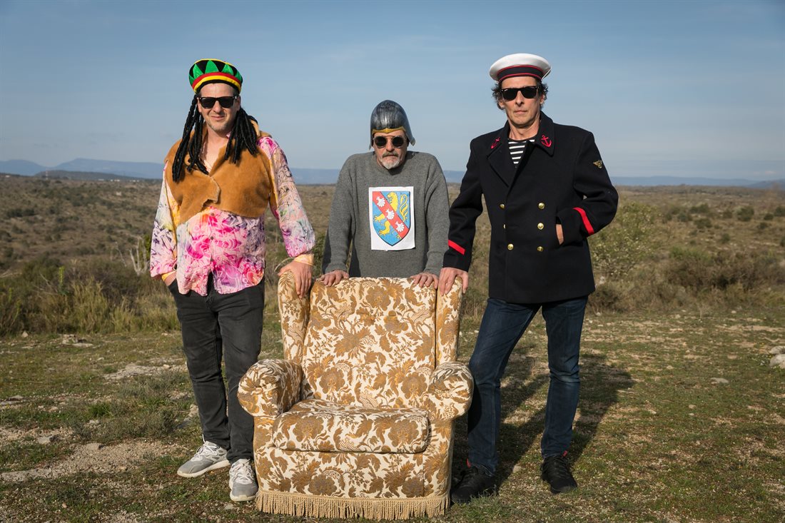 Groupe de musique Oscar m'a dit pose déguisés, derrière et mains sur fauteuil vintage en tissu à motifs floraux et à franges, paysage ardéchois en fond, montagnes, végétation pauvre, ambiance très lumineuse 