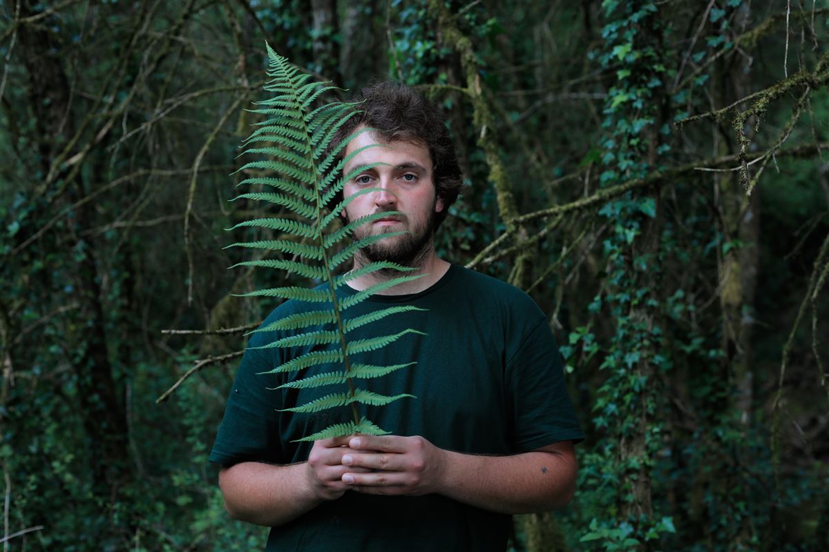 Léo en t-shirt vert dans son univers de verdure, il tient une fougère verte comme tout le vert naturel qui l'entoure.