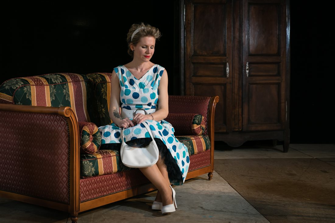 Décor de théâtre minimaliste, Eva assise sur canapé bois à l'ancienne vert et bordeaux, pose en robe vintage à pois bleus un sac à main blanc dans les mains, regard pudique vers le sol, vieille armoire en fond  