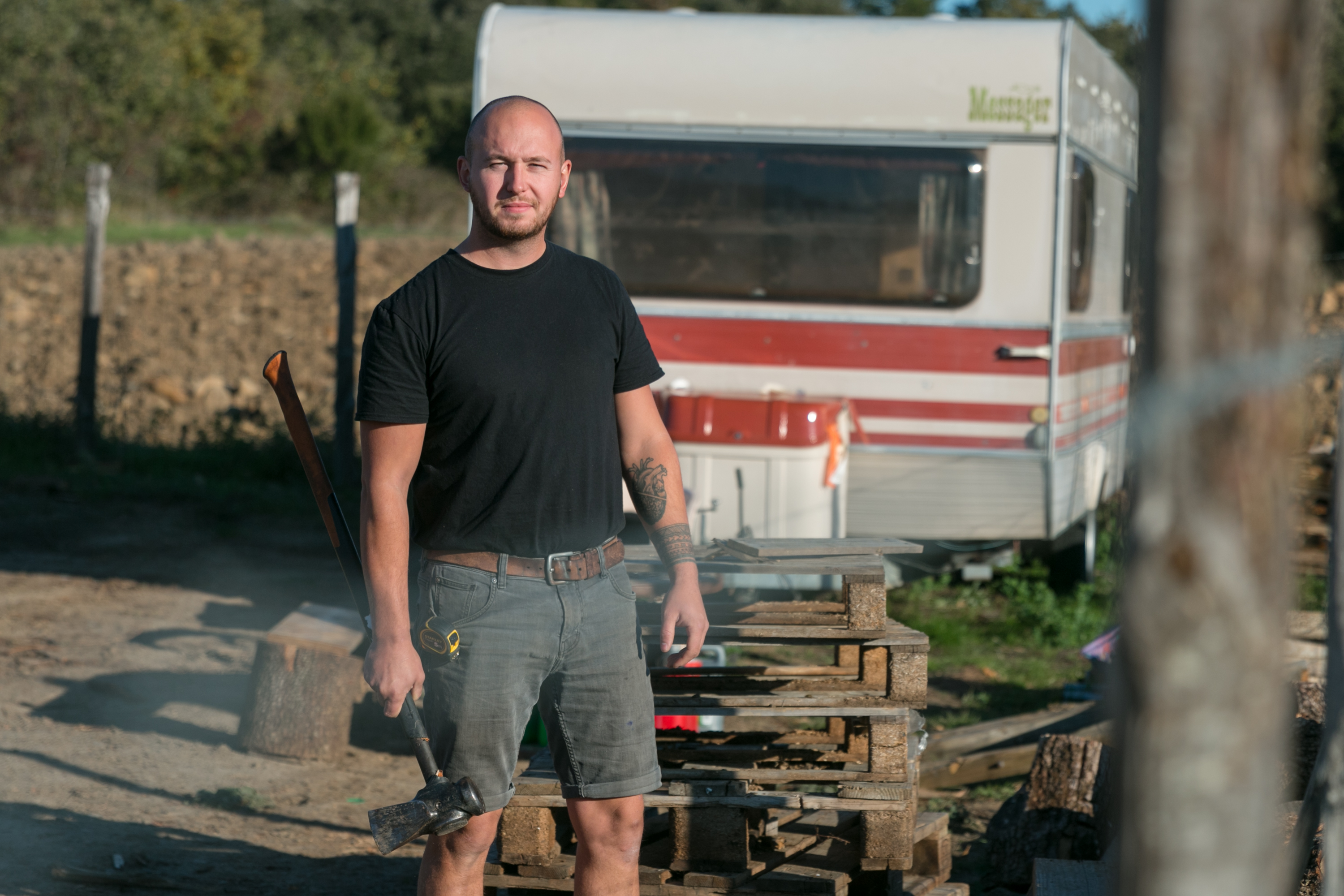 Piere, debout, jeune bûcheron, merlin à fendre dans la main droite, en fond caravane refuge pour repos, photo très lumineuse