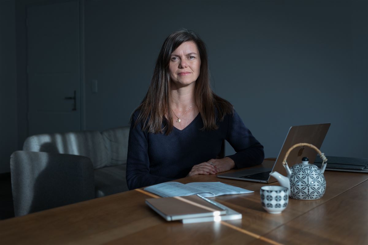 Véronique chez elle, assise devant son ordinateur de Web-rédactrice, le bras gauche posé sur le bureau, une théière et un tasse à portée de main.